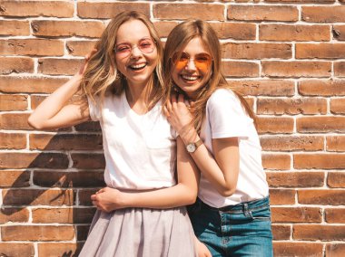 Trendy yaz beyaz t-shirt giysiler iki genç güzel sarışın gülümseyen hipster kız. Tuğla duvarın yanındaki sokakta poz veren kadınlar. Güneş gözlüğü ile eğlenen pozitif modeller 