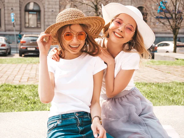 トレンディな夏の白いTシャツの服を着た2人の若い美しいブロンドの笑顔のヒップスターの女の子の肖像画 ストリートの背景にポーズセクシーな気楽な女性 サングラスと帽子で楽しむポジティブなモデル — ストック写真