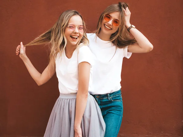 トレンディな夏の白いTシャツの服を着た2人の若い美しいブロンドの笑顔ヒップスターの女の子 赤い壁の近くの通りでポーズをとる女性たち サングラスで楽しむポジティブなモデル — ストック写真