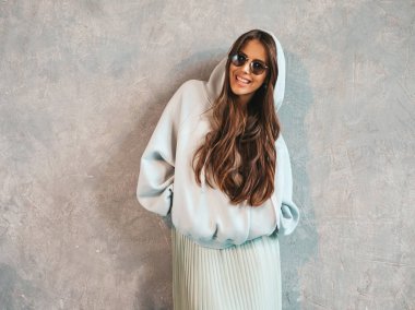 Genç, güzel, gülümseyen bir kadın kameraya bakıyor. Gündelik kapüşonlu ve etekli moda kız. Stüdyoda gri duvarın yanında poz veren komik ve pozitif bir kadın. Güneş gözlükleriyle