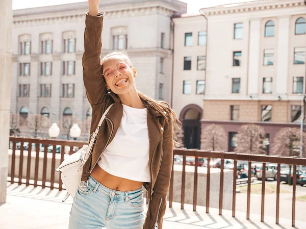 Portrait of beautiful smiling brunette model dressed in summer hipster jacket clothes. Trendy girl posing in the street background. Funny and positive woman in round sunglasses.Raising her hand