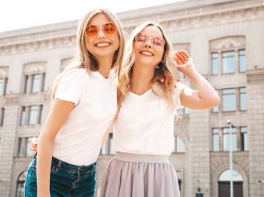 Moda yaz beyaz t-shirt giysiler iki genç güzel sarışın gülümseyen hipster kız Portresi. Seksi kaygısız kadınlar sokak arka planda poz. Güneş gözlüğü ile eğlenen pozitif modeller. Sarılma