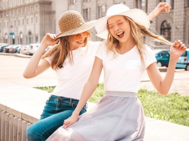 Moda yaz beyaz t-shirt giysiler iki genç güzel sarışın gülümseyen hipster kız Portresi. Seksi kaygısız kadınlar sokak arka planda poz. Güneş gözlüğü ve şapka ile eğlenen pozitif modeller