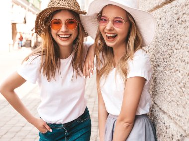 Trendy yaz beyaz t-shirt giysiler iki genç güzel sarışın gülümseyen hipster kız. Duvarın yanındaki sokakta poz veren kadınlar. Güneş gözlüğü ve şapka ile eğlenen pozitif modeller