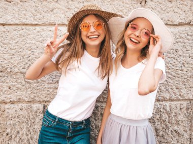 Trendy yaz beyaz t-shirt giysiler iki genç güzel sarışın gülümseyen hipster kız. Duvarın yanındaki sokakta poz veren kadınlar. Güneş gözlüğü ve şapka ile eğlenen pozitif modeller