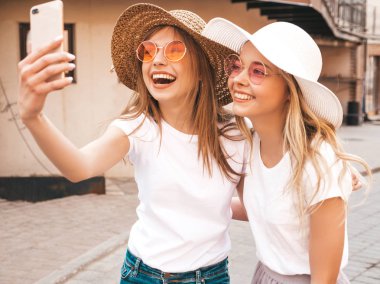 İki genç, gülümseyen, beyaz tişörtlü sarışın kadın. Akıllı telefondan selfie çeken kızlar. Arka planda poz veren mankenler. Olumlu yüz ifadeleri gösteren bir kadın.