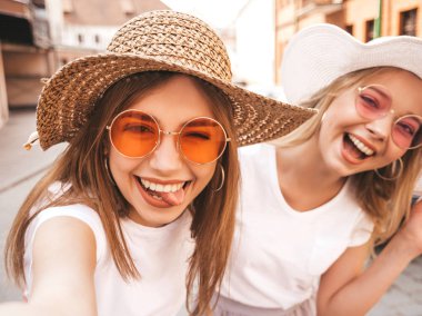 Yaz beyaz t-shirt iki genç gülümseyen hipster sarışın kadın. Akıllı telefonda selfie selfie portre fotoğrafları çeken kızlar. Sokak arka planında poz veren mankenler. Kadın dil ve olumlu duygular gösterir