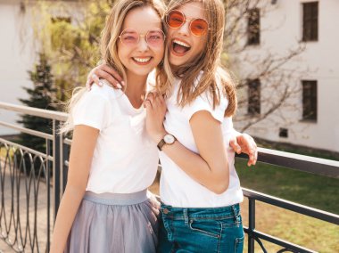 Moda yaz beyaz t-shirt giysiler iki genç güzel sarışın gülümseyen hipster kız Portresi. Seksi kaygısız kadınlar sokak arka planda poz. Güneş gözlüğü ile eğlenen pozitif modeller. Sarılma