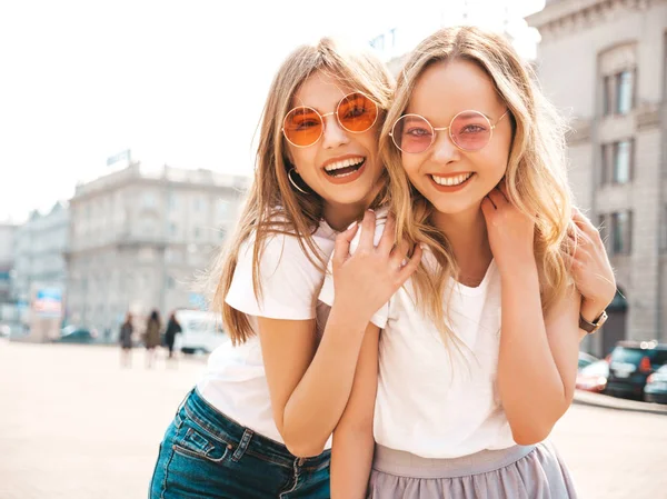 Moda yaz beyaz t-shirt giysiler iki genç güzel sarışın gülümseyen hipster kız Portresi. Seksi kaygısız kadınlar sokak arka planda poz. Güneş gözlüğü ile eğlenen pozitif modeller. Sarılma