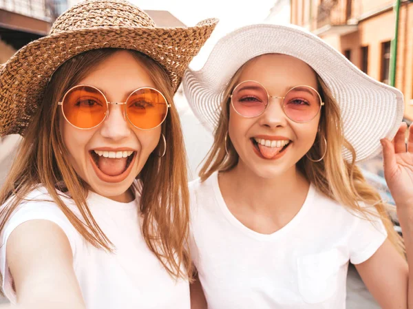 两个年轻的微笑的嬉皮士金发女郎在夏天的白色T恤 女孩在智能手机上自拍自拍照片 模特在街头背景上摆姿势 女性表现出舌头和积极情绪 — 图库照片