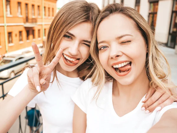 两个年轻微笑的时髦金发女郎穿着夏天的白色T恤 女孩在智能手机上自拍自拍照片 模特在街头背景上摆姿势 女性显示和平标志和舌头 — 图库照片