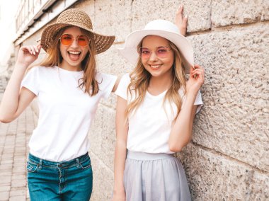 Moda yaz beyaz t-shirt giysiler iki genç güzel sarışın gülümseyen hipster kız Portresi. Seksi kaygısız kadınlar sokak arka planda poz. Güneş gözlüğü ve şapka ile eğlenen pozitif modeller