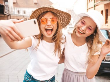 Yaz beyaz t-shirt iki genç gülümseyen hipster sarışın kadın. Akıllı telefonda selfie selfie portre fotoğrafları çeken kızlar. Sokak arka planında poz veren mankenler. Kadın dil ve olumlu duygular gösterir