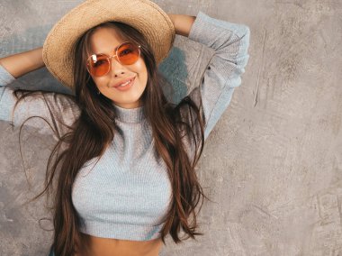 Genç, güzel, gülümseyen bir kadın kameraya bakıyor. Sıradan yaz kıyafetleri ve şapkası olan modaya uygun bir kız. Stüdyoda gri duvarın yanında poz veren komik ve pozitif kadın.