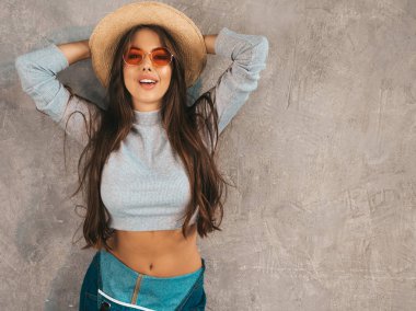 Genç, güzel, gülümseyen bir kadın kameraya bakıyor. Sıradan yaz kıyafetleri ve şapkası olan modaya uygun bir kız. Stüdyoda gri duvarın yanında poz veren komik ve pozitif kadın.
