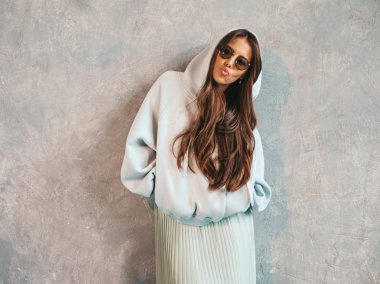 Genç, güzel, gülümseyen bir kadın kameraya bakıyor. Gündelik kapüşonlu ve etekli moda kız. Stüdyoda gri duvarın yanında poz veren komik ve pozitif bir kadın. Güneş gözlükleriyle. Ördek suratlı.