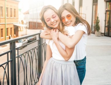 Moda yaz beyaz t-shirt giysiler iki genç güzel sarışın gülümseyen hipster kız Portresi. Seksi kaygısız kadınlar sokak arka planda poz. Güneş gözlüğü ile eğlenen pozitif modeller