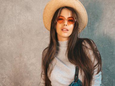 Kameraya bakan Genç güzel kadının portresi. Rahat yaz tulum giysi ve şapka Trendy kız. Komik ve pozitif kadın güneş gözlüğü stüdyoda gri duvar yakınında poz