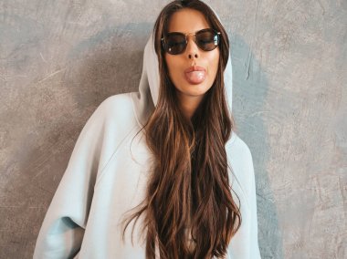 Young beautiful smiling woman looking at camera. Trendy girl in casual summer hoodie and skirt clothes. Funny and positive female posing near gray wall in studio in sunglasses.Shows tongue