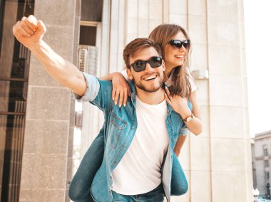 Gülümseyen güzel kız ve rahat yazlık giysiler onun yakışıklı erkek arkadaşı. Kız arkadaşını sırtında taşıyan adam ellerini kaldırıyor. Mutlu neşeli aile sokak arka planda eğlenmek 