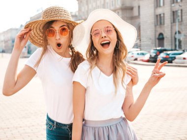 Trendy yaz beyaz t-shirt giysiler iki genç güzel sarışın gülümseyen hipster kız. Sokakta poz veren seksi şok kadınlar. Güneş gözlüğü ve şapka ile eğlenen sürpriz modeller. Barış işaretini gösterir