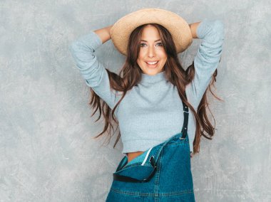 Kameraya bakan genç güzel kadının portresi. Rahat yaz tulum giysi ve şapka Trendy kız. Komik ve pozitif kadın stüdyoda gri duvar yakınında poz