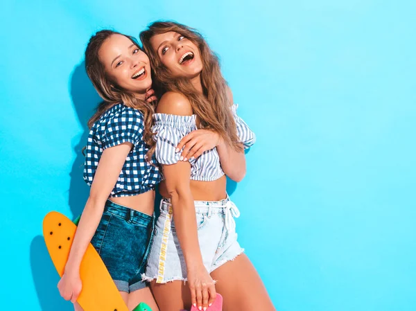 两个年轻的时尚微笑美丽的女孩与五颜六色的便士滑板 女人在夏天嬉皮士格子衬衫衣服摆在蓝色墙壁附近的工作室 积极的模型有乐趣和疯狂 — 图库照片