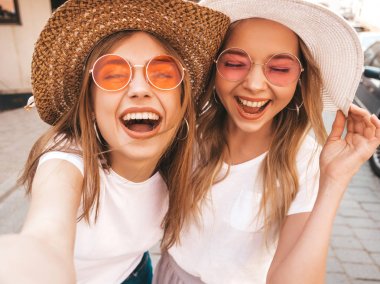 Yaz beyaz t-shirt iki genç gülümseyen hipster sarışın kadın. Akıllı telefonda selfie selfie portre fotoğrafları çeken kızlar. Sokak arka planında poz veren mankenler. Kadın dil ve olumlu duygular gösterir