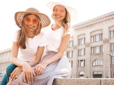 Moda yaz beyaz t-shirt giysiler iki genç güzel sarışın gülümseyen hipster kız Portresi. Seksi kaygısız kadınlar sokak arka planda poz. Güneş gözlüğü ve şapka ile eğlenen pozitif modeller