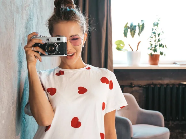 レトロなカメラで写真を撮る若い笑顔の写真家の女の子 女性が写真を撮る カジュアルな夏の白いTシャツに身を包んだモデル 灰色の壁の近くのスタジオでポーズ — ストック写真