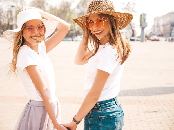 两个年轻美丽的金发微笑的嬉皮士女孩在时髦的夏季白色T恤衣服和帽子的背面 性感的无忧无虑的女人在街头背景摆姿势 情侣互相牵手 — 图库照片