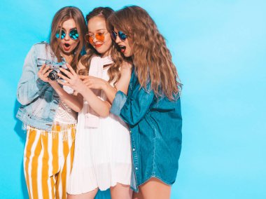 Trendy yaz rahat giysiler ve güneş gözlüğü üç genç güzel gülümseyen hipster kız. Seksi kaygısız kadınlar mavi duvarın yakınında poz. Retro kamera da yapılan resimlere bakmak