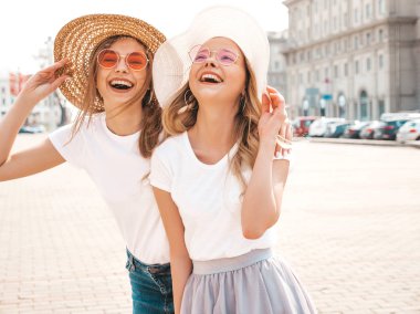 Moda yaz beyaz t-shirt giysiler iki genç güzel sarışın gülümseyen hipster kız Portresi. Seksi kaygısız kadınlar sokak arka planda poz. Güneş gözlüğü ve şapka ile eğlenen pozitif modeller