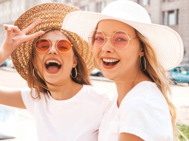 Moda yaz beyaz t-shirt giysiler iki genç güzel sarışın gülümseyen hipster kız Portresi. Seksi kaygısız kadınlar sokak arka planda poz. Güneş gözlüğü ve şapka ile eğlenen pozitif modeller
