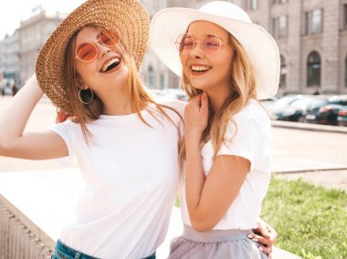 Moda yaz beyaz t-shirt giysiler iki genç güzel sarışın gülümseyen hipster kız Portresi. Seksi kaygısız kadınlar sokak arka planda poz. Güneş gözlüğü ve şapka ile eğlenen pozitif modeller