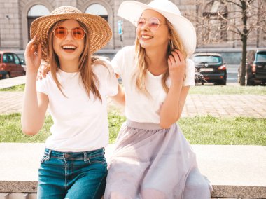 Moda yaz beyaz t-shirt giysiler iki genç güzel sarışın gülümseyen hipster kız Portresi. Seksi kaygısız kadınlar sokak arka planda poz. Güneş gözlüğü ve şapka ile eğlenen pozitif modeller