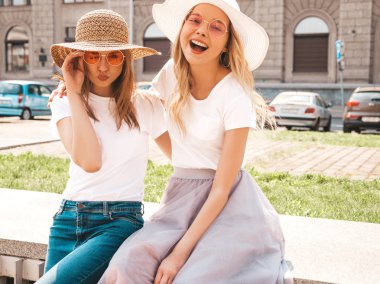 Moda yaz beyaz t-shirt giysiler iki genç güzel sarışın gülümseyen hipster kız Portresi. Seksi kaygısız kadınlar sokak arka planda poz. Güneş gözlüğü ve şapka ile eğlenen pozitif modeller