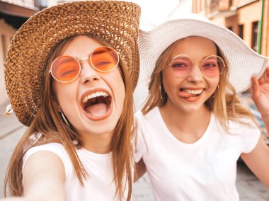 Yaz beyaz t-shirt iki genç gülümseyen hipster sarışın kadın. Akıllı telefonda selfie selfie portre fotoğrafları çeken kızlar. Sokak arka planında poz veren mankenler. Kadın olumlu duygular gösterir