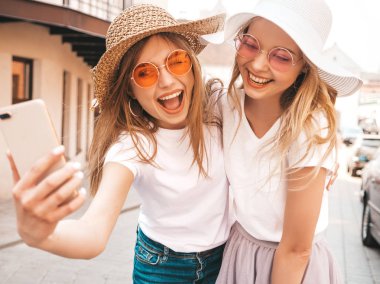 Yaz beyaz t-shirt iki genç gülümseyen hipster sarışın kadın. Akıllı telefonda selfie selfie portre fotoğrafları çeken kızlar. Sokak arka planında poz veren mankenler. Kadın olumlu duygular gösterir