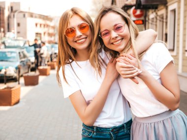 Moda yaz beyaz t-shirt giysiler iki genç güzel sarışın gülümseyen hipster kız Portresi. Seksi kaygısız kadınlar sokak arka planda poz. Güneş gözlüğü ile eğlenen pozitif modeller. Sarılma