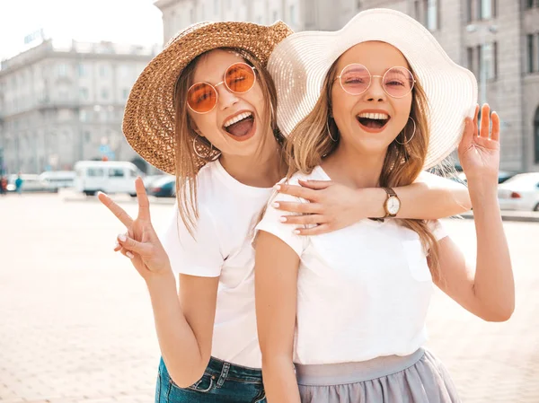两个年轻美丽的金发微笑的嬉皮士女孩在时髦的夏季白色T恤衣服的肖像 性感的无忧无虑的女人在街头背景摆姿势 正款车型在太阳镜和帽子的乐趣 — 图库照片