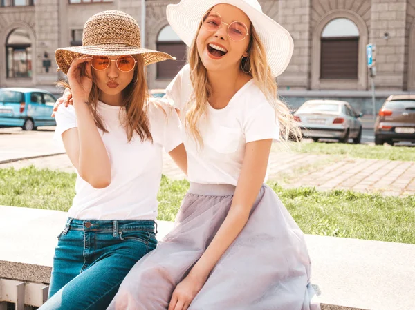 トレンディな夏の白いTシャツの服を着た2人の若い美しいブロンドの笑顔のヒップスターの女の子の肖像画 ストリートの背景にポーズセクシーな気楽な女性 サングラスと帽子で楽しむポジティブなモデル — ストック写真