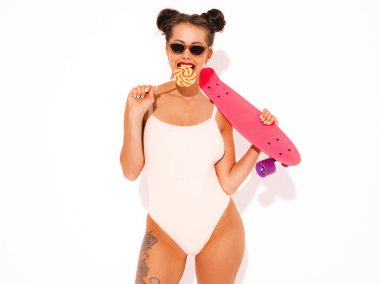 Güneş gözlüğü kırmızı dudaklar ile Genç güzel seksi gülümseyen hipster kadın. Yazlık mayo giyen kız. Pembe peni kaykay ile deli gidiyor, beyaz izole. İki boynuz. Şeker lolipop yeme