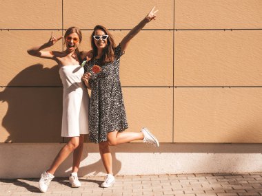 Trendy yaz elbisesi iki genç güzel gülümseyen hipster kız. Seksi kaygısız kadınlar sarı duvarın yakınındaki sokakta poz. Güneş gözlüğü ile eğlenen pozitif modeller. Şekerli lolipop yiyorum. El kaldırma