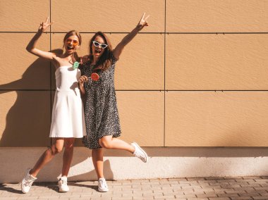 Trendy yaz elbisesi iki genç güzel gülümseyen hipster kız. Seksi kaygısız kadınlar sarı duvarın yakınındaki sokakta poz. Güneş gözlüğü ile eğlenen pozitif modeller. Şekerli lolipop yiyorum. El kaldırma