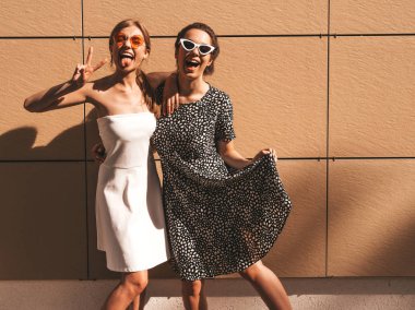 Trendy yaz elbisesi iki genç güzel gülümseyen hipster kız. Seksi kaygısız kadınlar sarı duvarın yakınındaki sokakta poz. Güneş gözlüğü ile eğlenen pozitif modeller. Sarılma, dil ve barış işareti gösterir