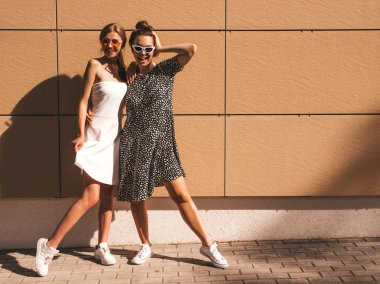 Trendy yaz elbisesi iki genç güzel gülümseyen hipster kız. Seksi kaygısız kadınlar sarı duvarın yakınındaki sokakta poz. Güneş gözlüğü ile eğlenen pozitif modeller. Sarılma. Tam uzunlukta