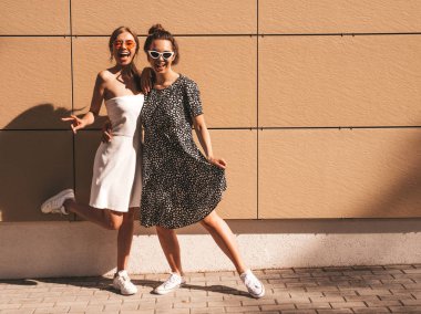 Trendy yaz elbisesi iki genç güzel gülümseyen hipster kız. Seksi kaygısız kadınlar sarı duvarın yakınındaki sokakta poz. Güneş gözlüğü ile eğlenen pozitif modeller. Sarılma. Tam uzunlukta