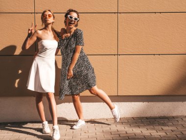 Trendy yaz elbisesi iki genç güzel gülümseyen hipster kız. Seksi kaygısız kadınlar sarı duvarın yakınındaki sokakta poz. Güneş gözlüğü ile eğlenen pozitif modeller. Sarılma. Tam uzunlukta