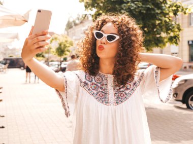 Yaz hipster beyaz elbise giymiş afro bukleler saç modeli ile güzel gülümseyen model. Seksi kaygısız kız güneş gözlüğü sokakta poz. Akıllı telefonda selfie selfie portre fotoğrafları çekmek. Ördek yüz
